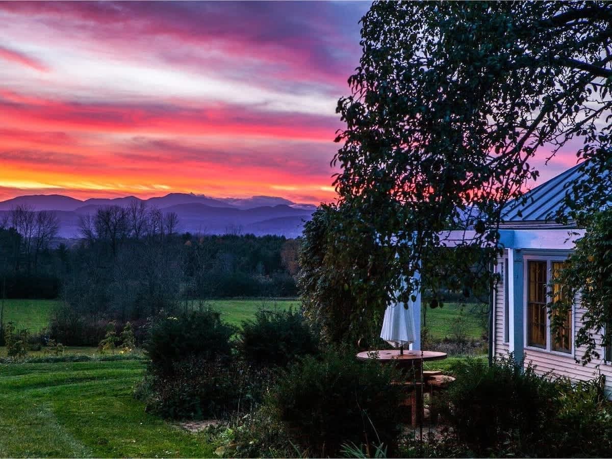 Colorful sunset over a rural landscape with mountains, trees, a house with windows, and a patio area with an umbrella.