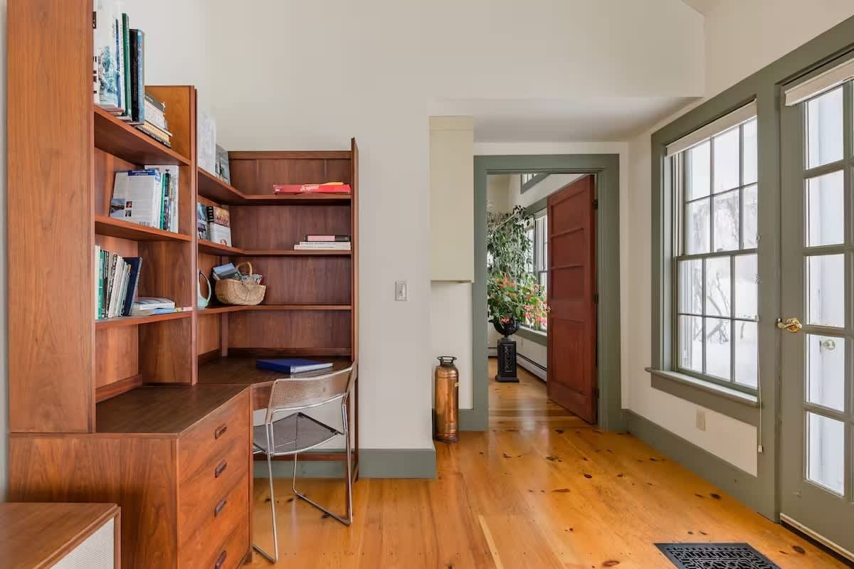 Interior view of a cozy room with a built-in wooden desk and bookshelves, hardwood floors, large windows, and a partially open door leading to a brightly lit space with plants.