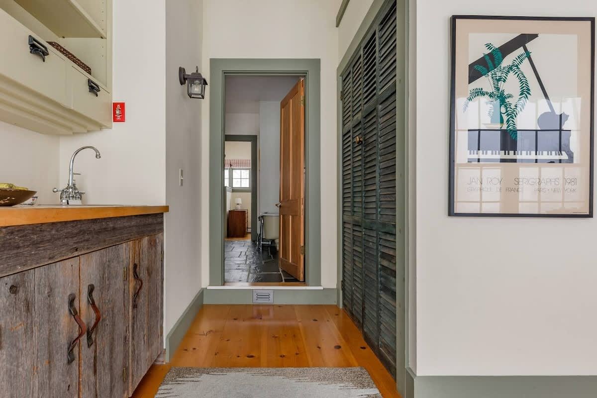 Hallway leading to a room with an open door, featuring a wooden cabinet with handles, a framed music poster on the wall, and a grey louvered closet door.