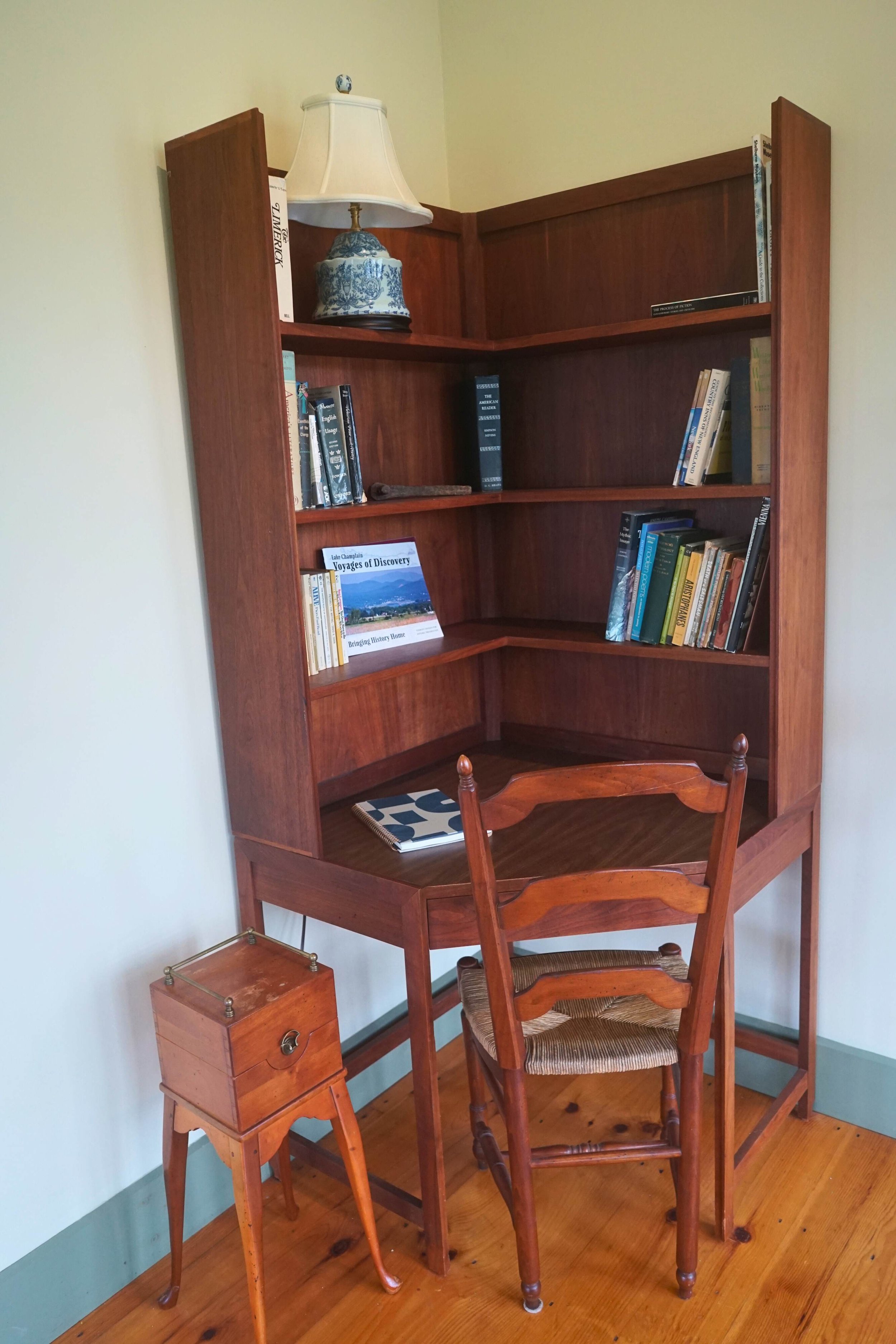 Wooden desk with shelves filled with books, a blue and white table lamp, a wooden chair, and a small wooden storage box on a tripod stand, against a light-colored wall.