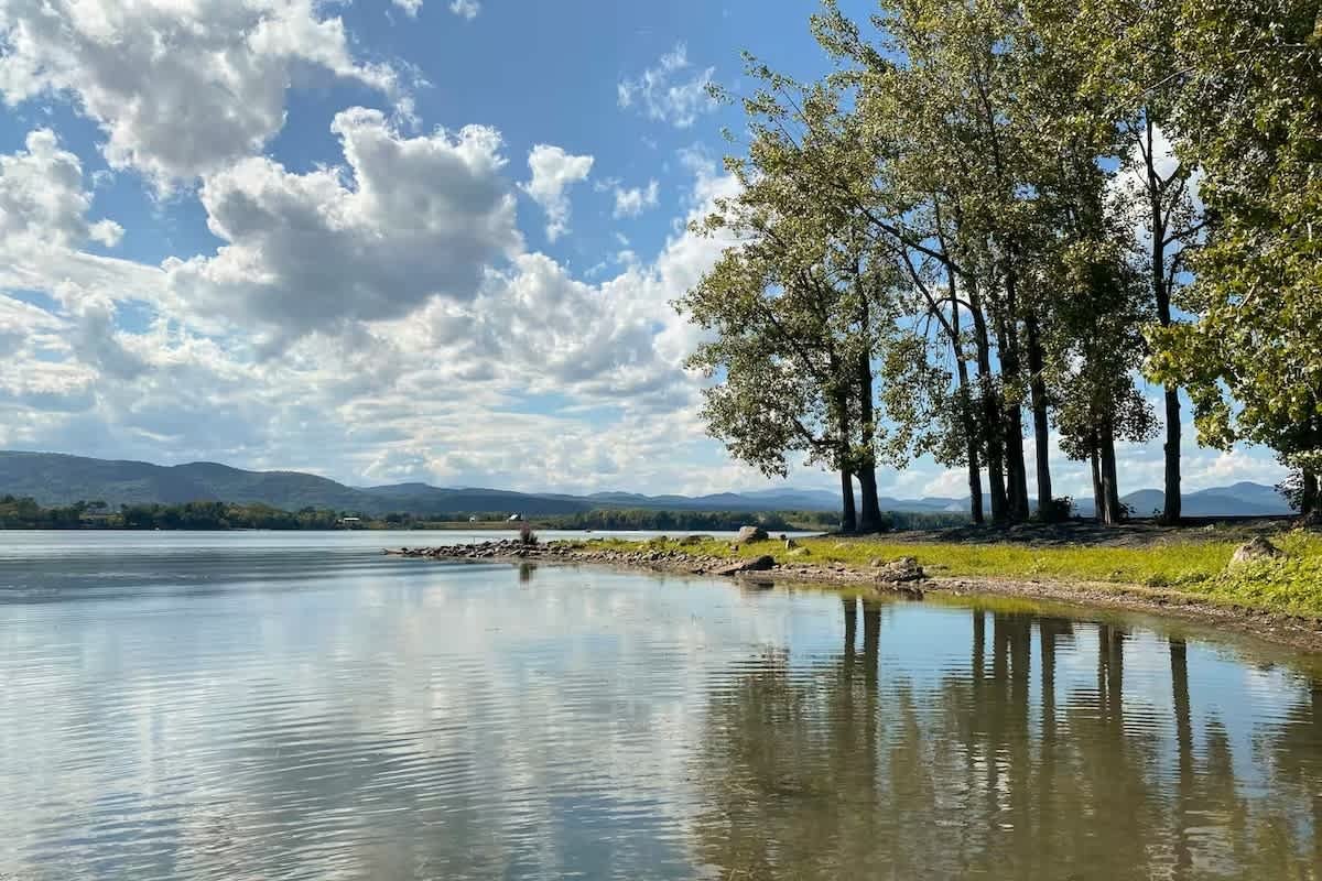 A calm lake with reflections of trees and a partly cloudy sky, with distant mountains on the horizon.