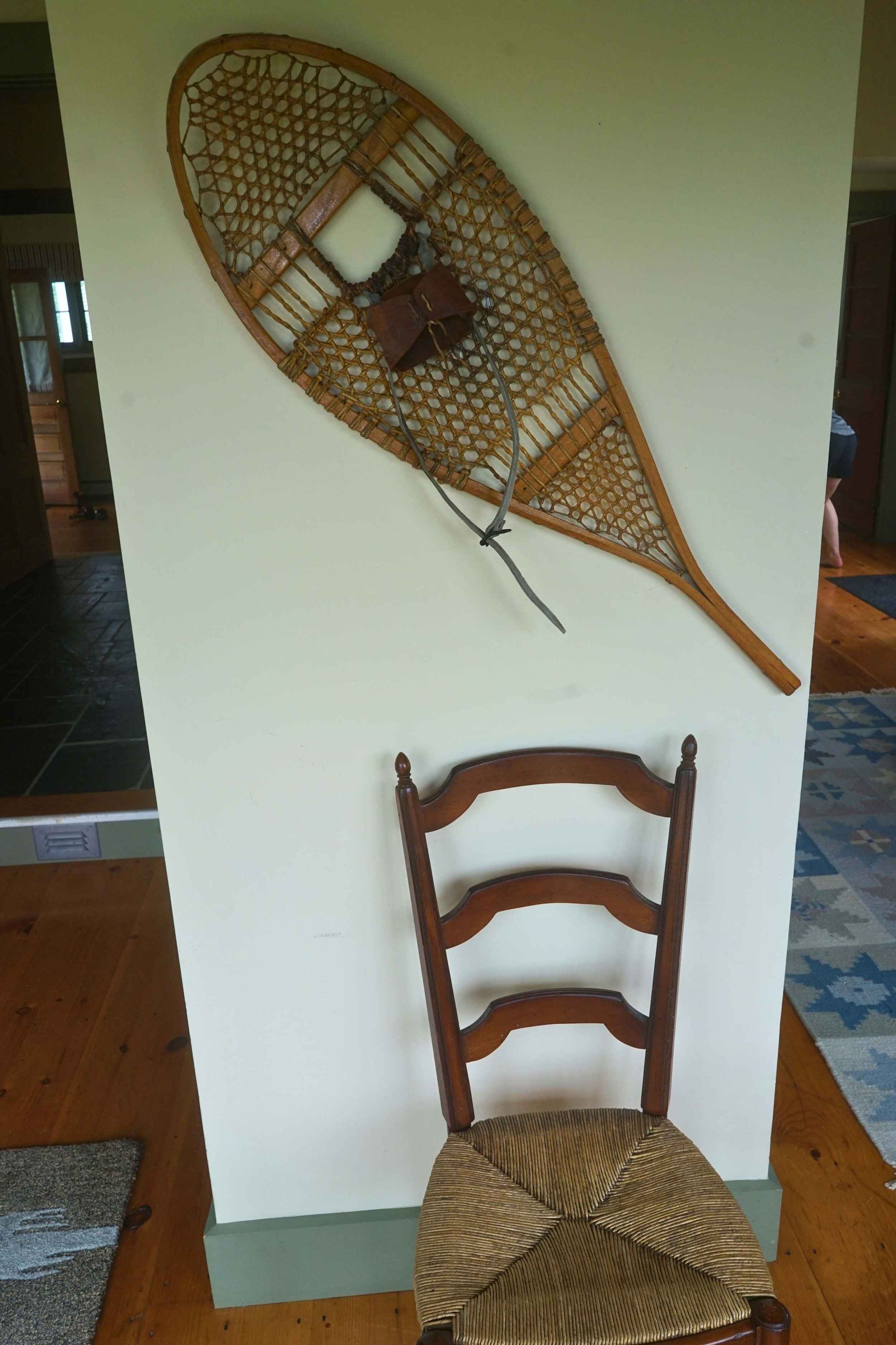 A vintage snowshoe mounted on a wall above a wooden chair with a woven seat.