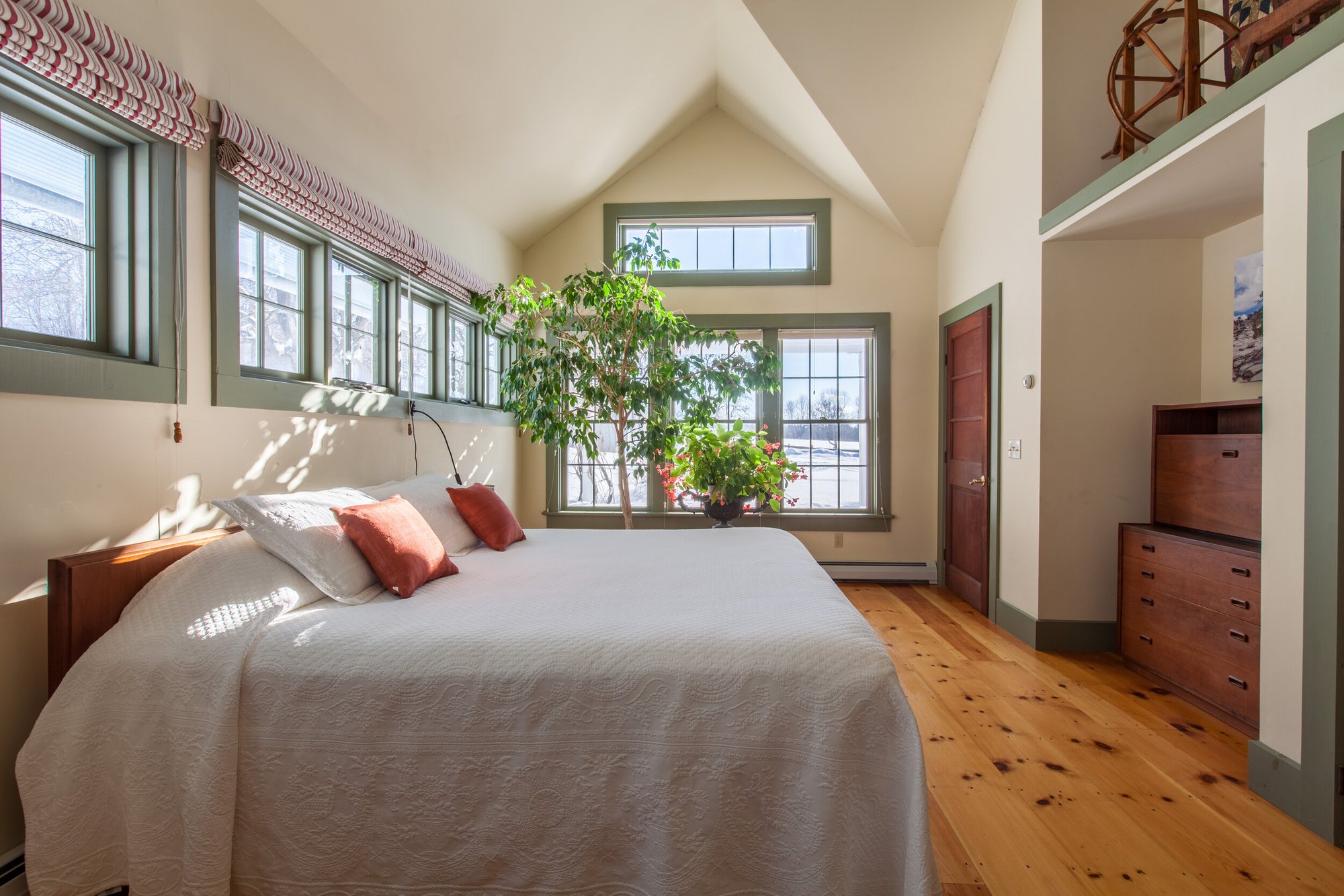 Guest house bedroom with vaulted ceiling, white bedding with orange accent pillows, wood floors, and large windows overlooking the Vermont countryside.