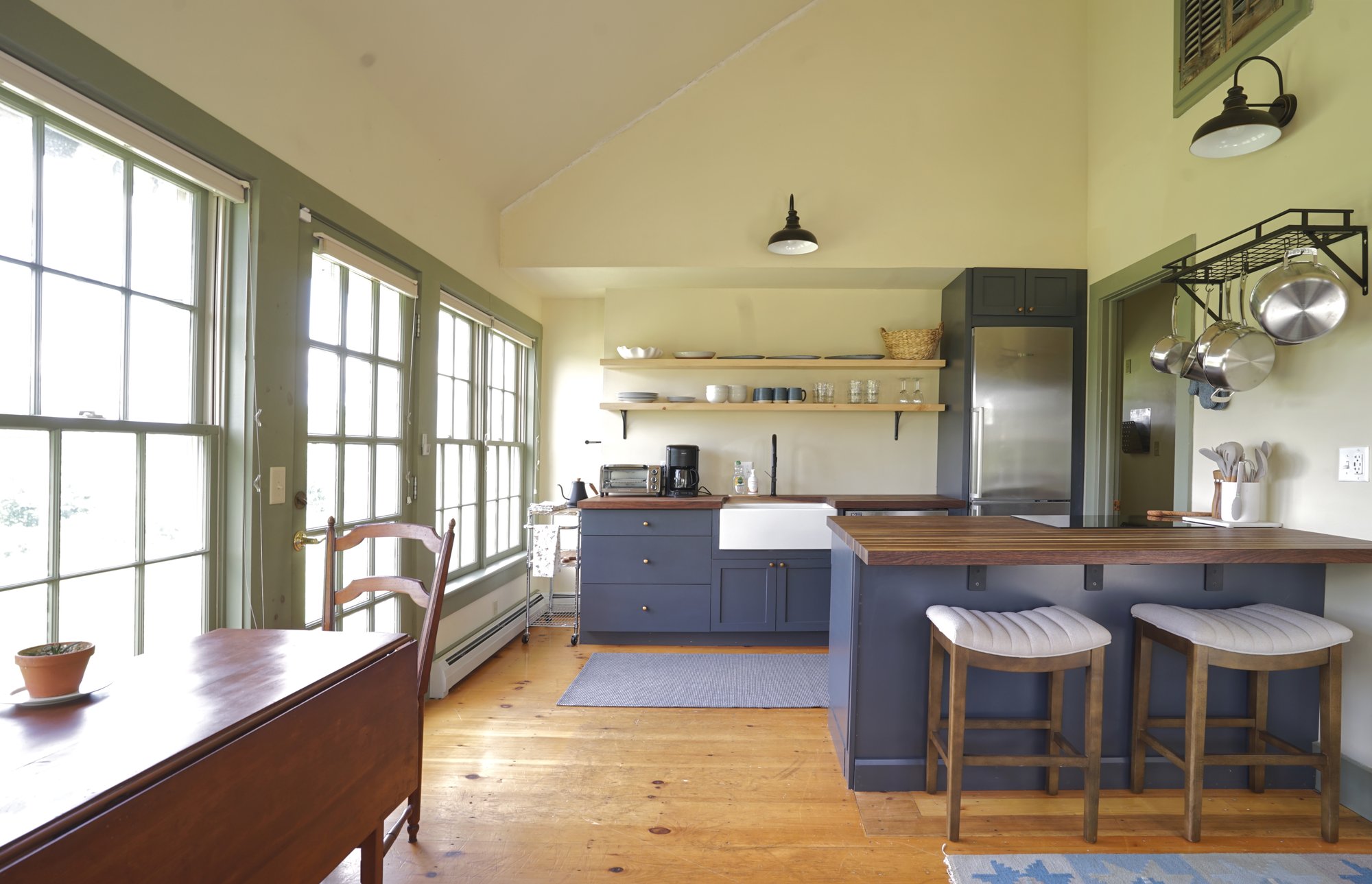 Sunlit guest house kitchen with blue cabinets, farmhouse sink, butcher-block island with two stools, and windows on all sides.