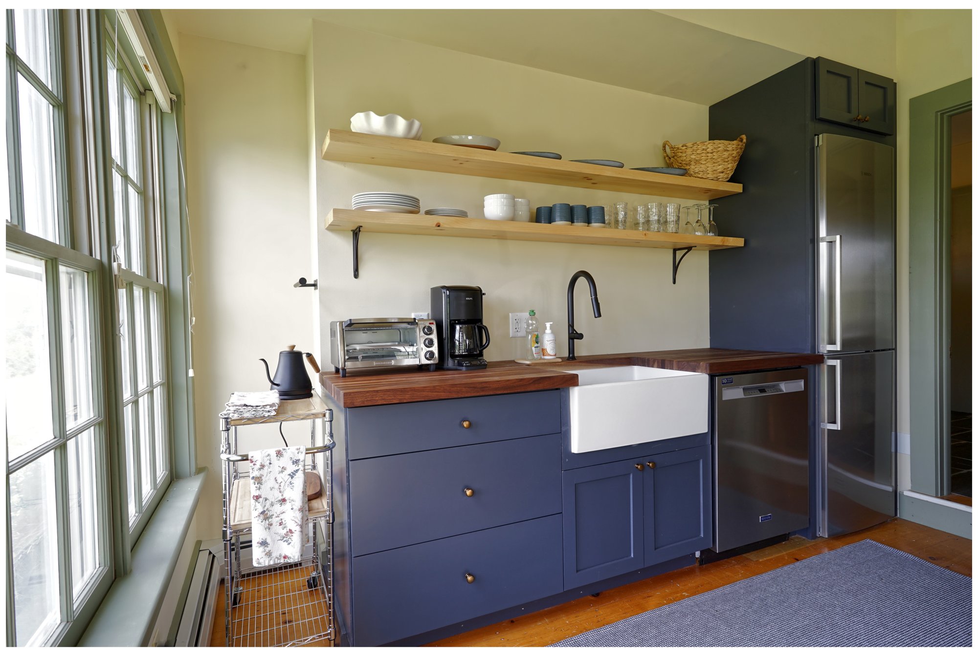 Kitchen with blue cabinets, farmhouse sink, open wooden shelves, and stainless refrigerator surrounded by bright windows.