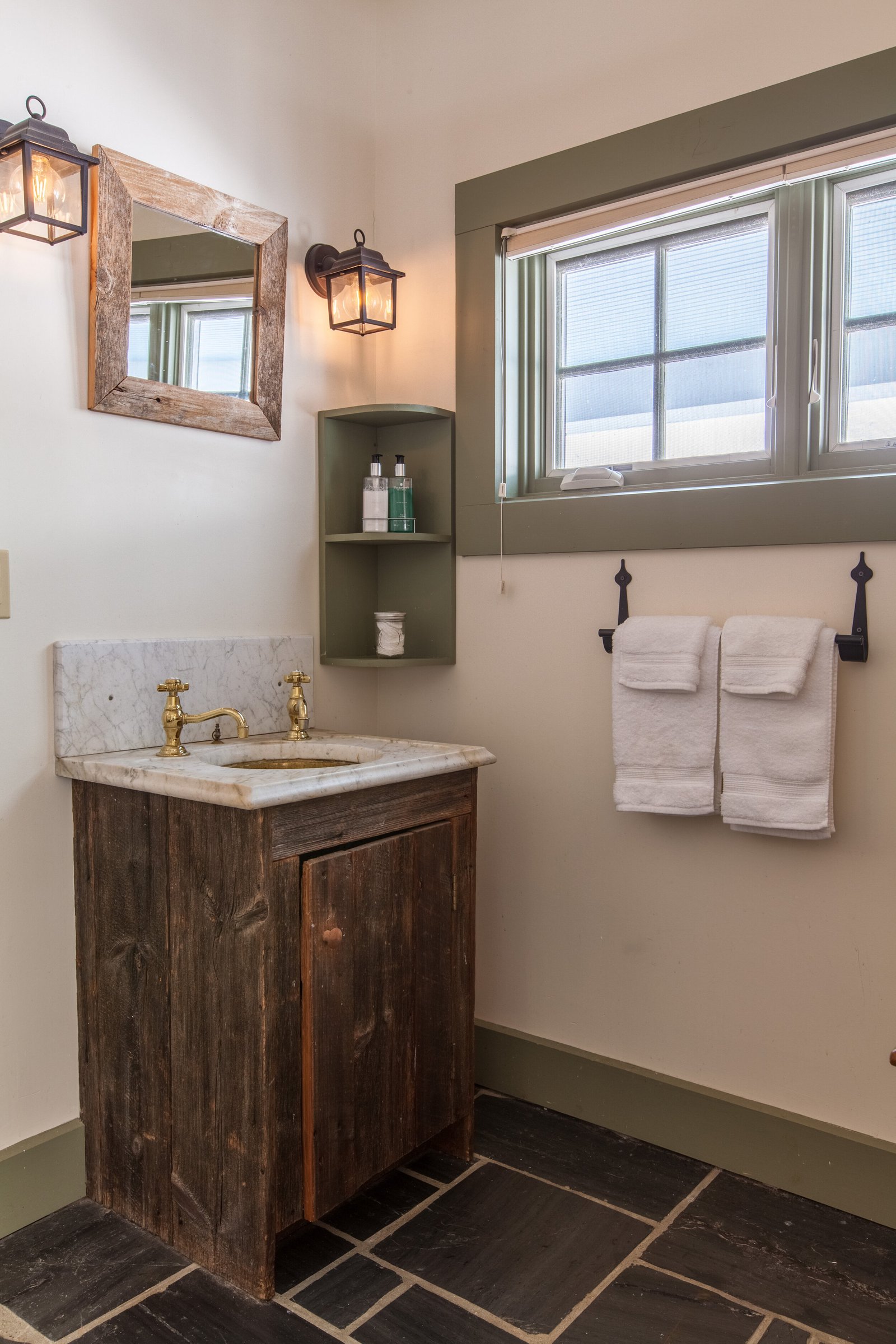 Rustic wood bathroom vanity with marble top, brass faucet, mirror, and a window with a countryside view.