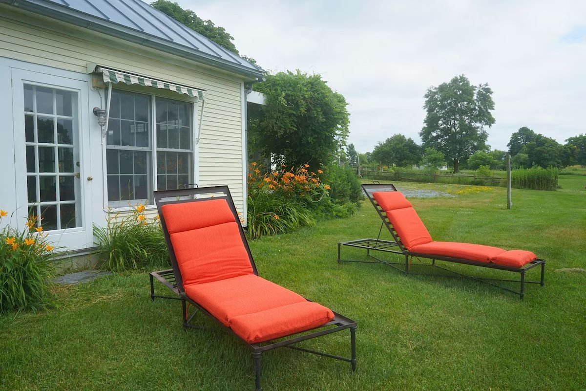 Two lounge chairs with orange cushions on the lawn beside the guest house, surrounded by daylilies and gardens.