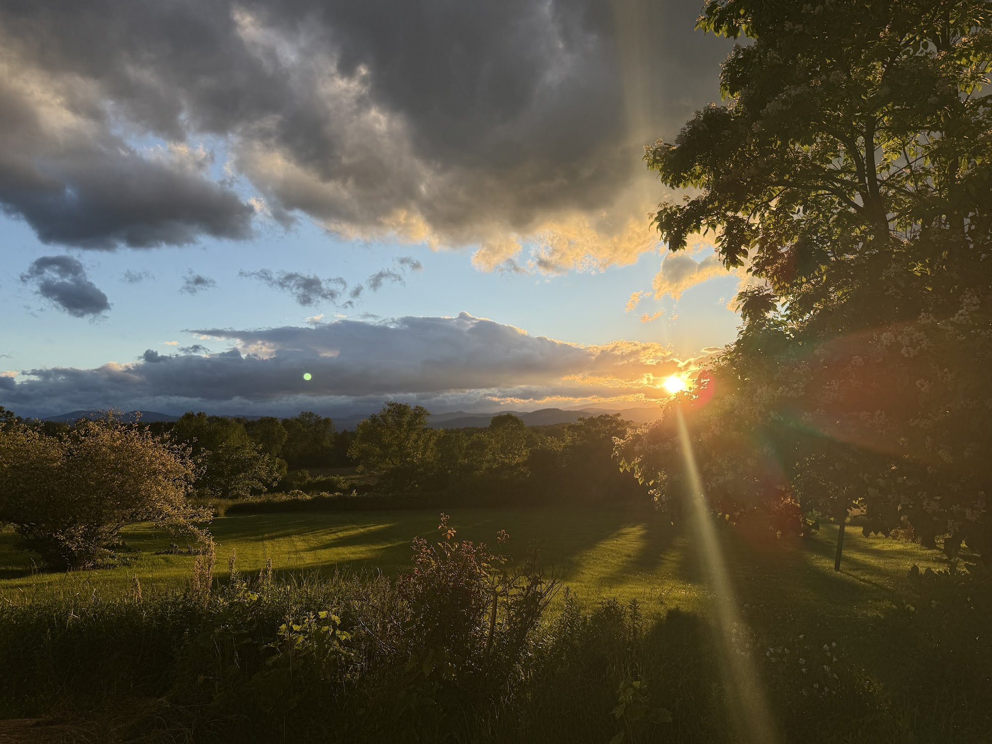 Sun setting over the fields beyond the guest house, with dramatic clouds and long shadows across the meadow.